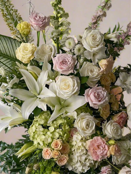 Close-up of white lilies and delicate blush pink roses in a funeral standing spray, arranged with lush green foliage in a church setting.