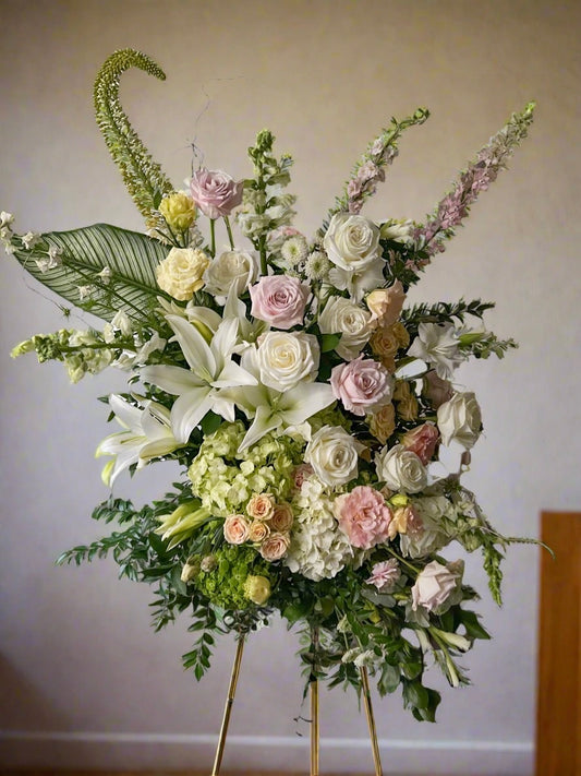 Exquisite funeral standing spray of white, green, and blush flowers in a church setting. The elegant arrangement features white lilies, roses, and soft blush pink blooms, complemented by lush green foliage. Positioned on a tall stand near the altar, the floral display creates a serene atmosphere with the church’s stained-glass windows and soft lighting in the background, offering a beautiful tribute for a memorial service.